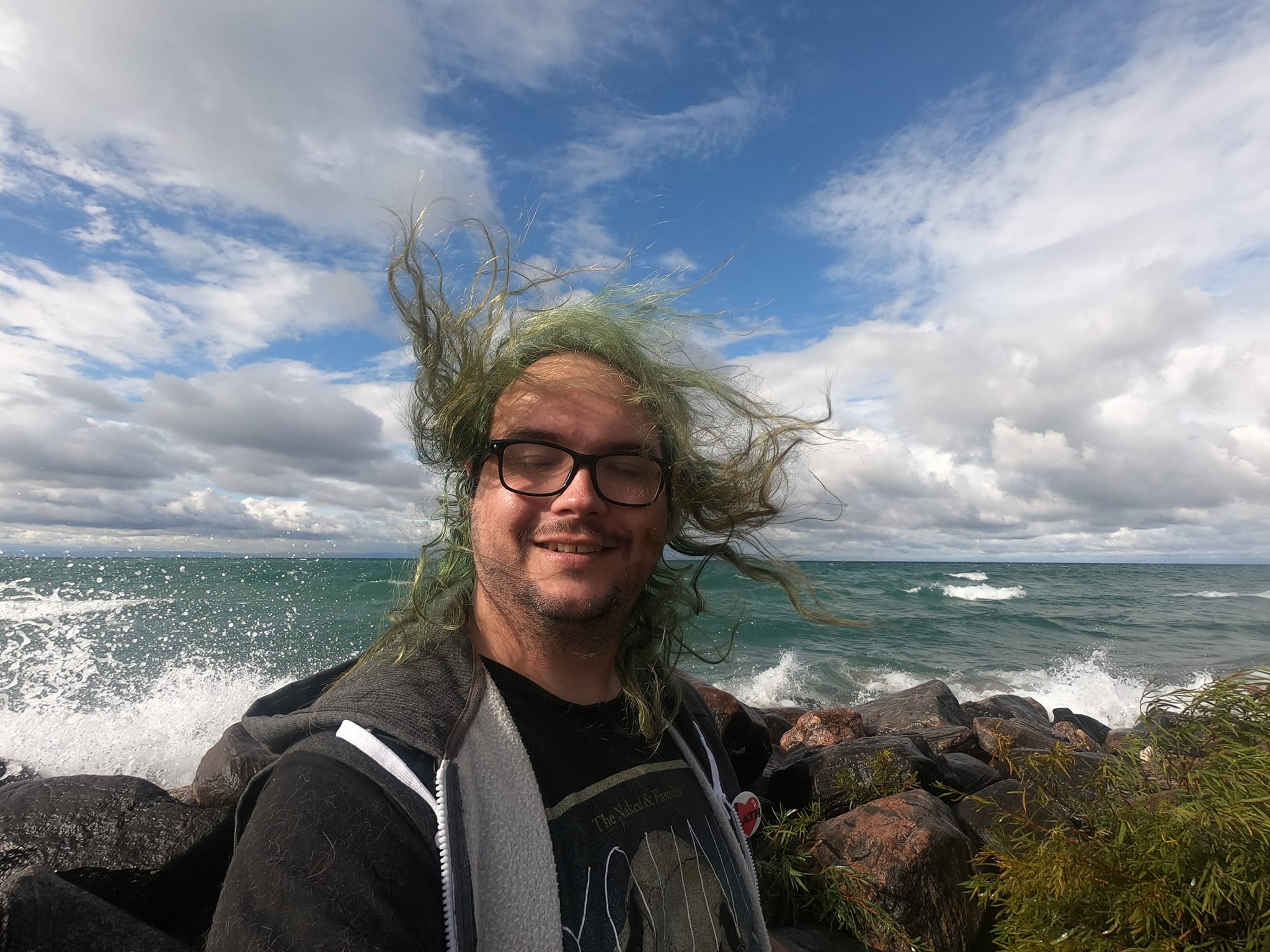 A man with long green hair wearing a hooded sweater vest stands at a breakwater, hair blowing in the wind, Photo 3